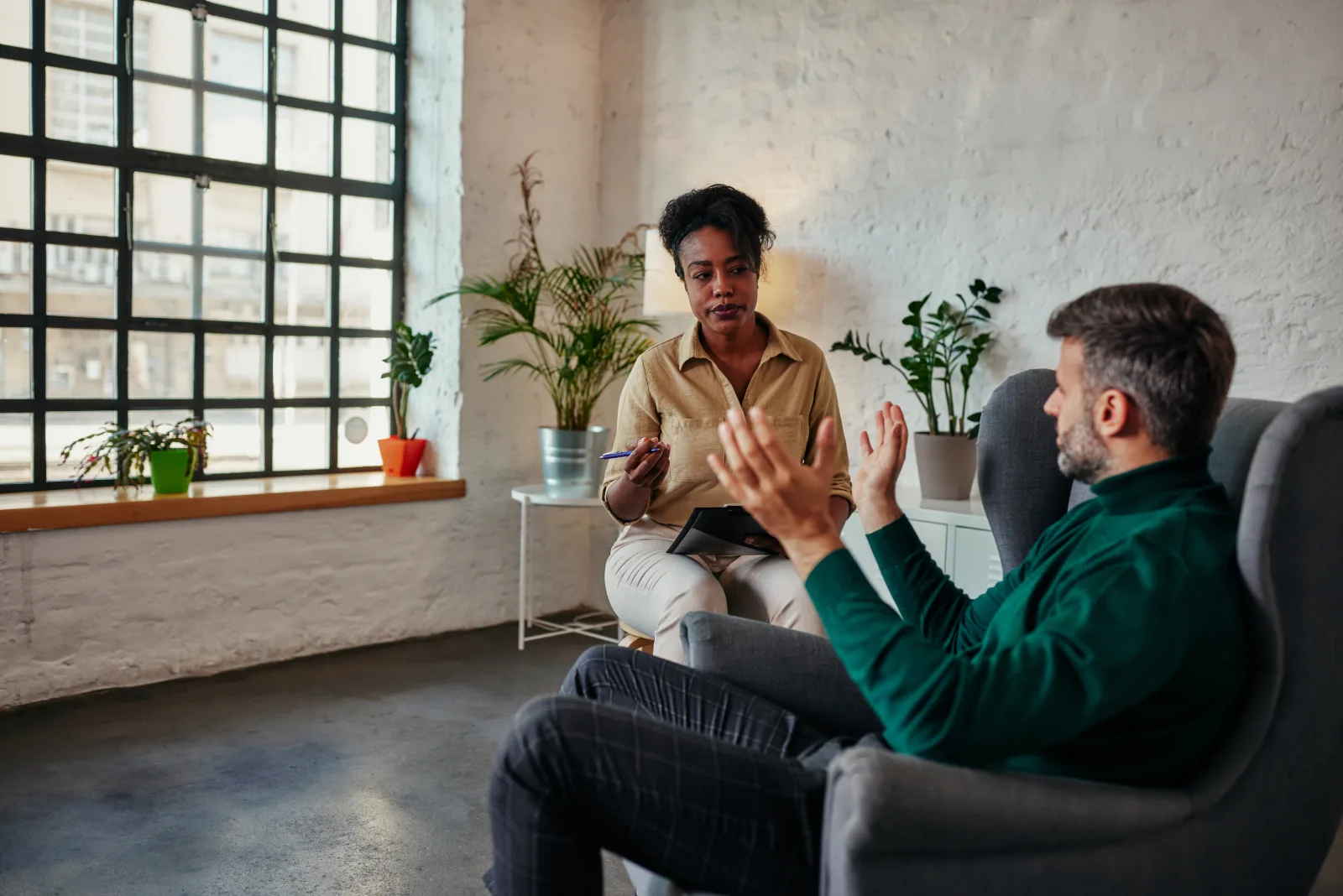 A therapist leaning in to talk with a client, the two seated facing each other in a bright, plant-filled room