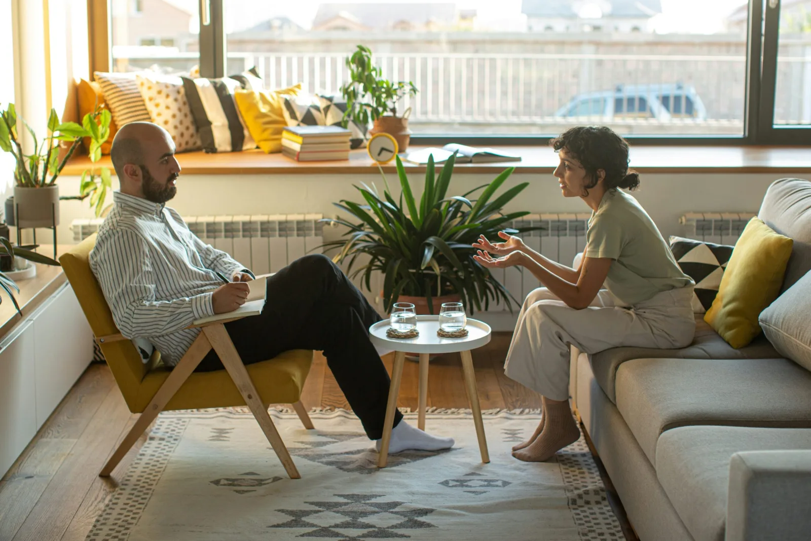 Two people mid-conversation in a sunlit, plant-filled living room, seated across a low table